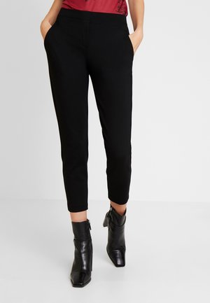 Femme portant un pantalon noir ajusté aux chevilles, des bottines noires à talons, et un haut rouge, debout les mains dans les poches.