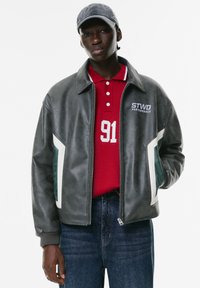 Giubbotto in ecopelle grigio con accenti bianchi e teal. Indossato sopra un polo rossa con colletto bianco e numero '91', abbinato a jeans in denim scuro.