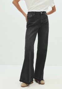 Femme portant un jean noir taille haute à jambes larges avec des ourlets bruts et des chaussures beiges, se tenant avec une main sur la hanche.