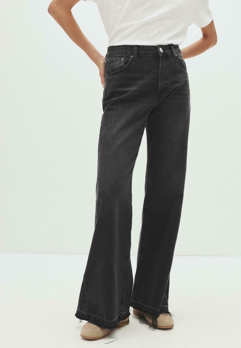 Femme portant un jean noir taille haute à jambes larges avec des ourlets bruts et des chaussures beiges, se tenant avec une main sur la hanche.