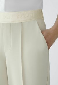Beige high-waisted trousers with a textured fabric, featuring a wide waistband embossed with "OUI" and side pockets.
