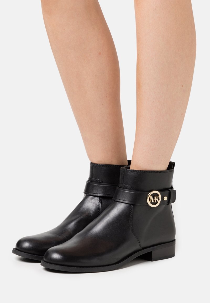 Jambes portant des bottines en cuir noir avec une sangle et une boucle circulaire dorée avec le logo MK sur le côté, sur fond blanc.