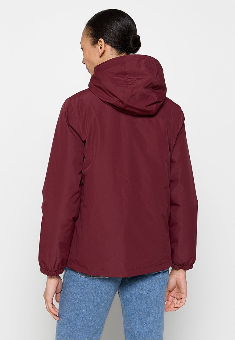 Chaqueta impermeable marrón con capucha, puños elásticos y una textura suave. Presenta un corte recto y está superpuesta sobre unos vaqueros azules.