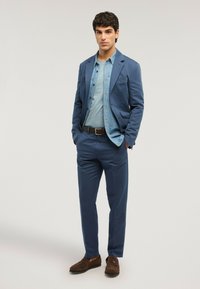 Joven de pie con las manos en los bolsillos, vistiendo un traje azul, camisa de mezclilla, camiseta azul clara, cinturón marrón y mocasines marrones.