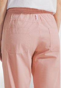 Jasnoróżowe spodnie z elastycznym paskiem w talii, dwoma tylnymi kieszeniami i materiałową metką z tyłu. Gładka tekstura i casualowy design.