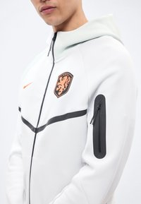 Νεαρό άτομο που φοράει λευκή ζακέτα με φερμουάρ, με πορτοκαλί έμβλημα λιονταριού στο στήθος και μαύρη τσέπη με φερμουάρ στο αριστερό μανίκι, μπροστά σε απλό φόντο.