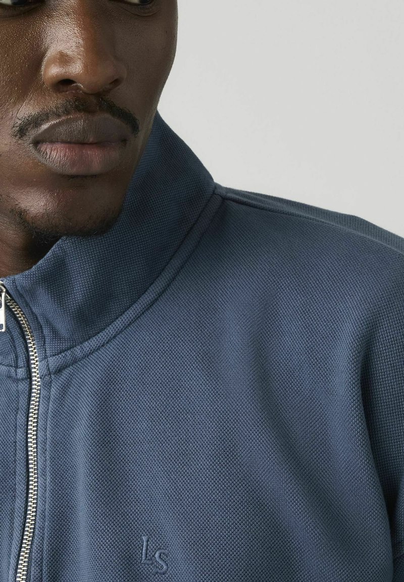 Primer plano de un hombre que lleva una chaqueta azul con cremallera y textura, con las letras "LS" bordadas en el pecho, mirando ligeramente hacia la izquierda sobre un fondo liso.
