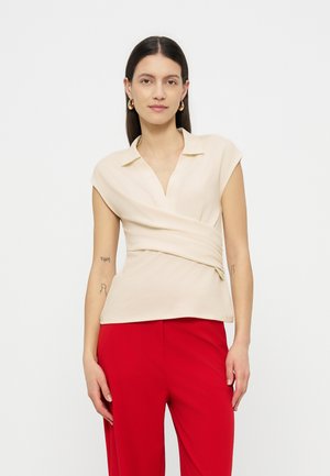 Žena s tamnom kosom nosi bež bezrukavu bluzu na preklop i crvene hlače, stoji ispred jednobojne svijetle pozadine.