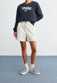 Nike mörkblå sweatshirt med logotyp, ljusbeige cargoshorts med elastisk midja och svartsprungna och vita rutiga slip-on sneakers.