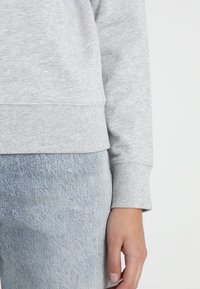 Felpa grigia chiara con orlo e polsi a costine, con una texture morbida. Indossata con jeans di denim blu chiaro, che mostrano un outfit casual.