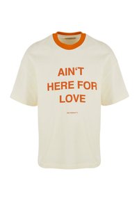 T-shirt en coton crème avec un col côtelé orange et un texte en gras : "JE NE SUIS PAS ICI POUR L'AMOUR" centré sur le devant. Coupe moderne, décontractée.
