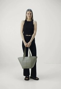 Kvinne står iført svart ermeløs topp og bukser, holder en stor grå totebag med begge hender foran seg.