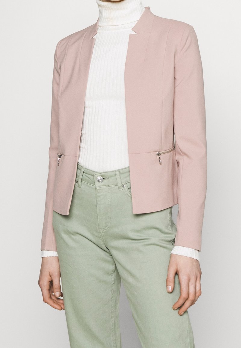 Personne portant un blazer rose clair avec des poches zippées à l'avant sur un col roulé côtelé blanc et un pantalon vert clair.