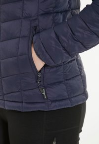 Marinblå pufferjacka med kviltad design, sidoficka med dragkedja och logotag på fållen. Slät, lättviktig material.