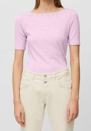 Frau trägt ein hellrosa kurzärmeliges Hemd und hochtaillierte cremefarbene Hosen, steht vor einem schlichten hellen Hintergrund.