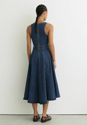 Sieviete ar pītām matām, tērpusies bezroku tumszili vidēja garuma džinsa kleitā ar rāvējslēdzēju mugurpusē, stāvot pretī baltai sienai.