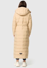 Manteau doudoune beige long avec capuche, présentant des sections matelassées, une taille ceinturée et des fermetures éclaire latérales. Porté sur un pantalon bleu et des baskets blanches.