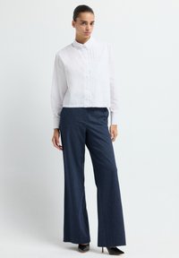Camicia bianca con colletto e bottoni sul davanti, abbinata a pantaloni larghi a righe navy e scarpe con tacco nere.