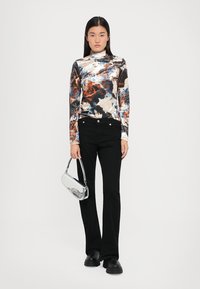 Langärmliges Turtleneck-Top mit buntem abstraktem Muster, kombiniert mit schwarzen Schlaghosen. Hält eine silberne Handtasche und trägt klobige schwarze Schuhe.