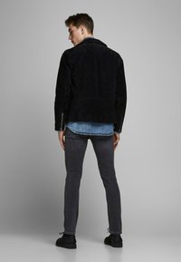 Schwarze Wildlederjacke mit Kragen, Rückennahtdetails, kombiniert mit einem Denim-Hemd und dunkelgrauen Slim-Fit-Jeans. Schwarze Schuhe.