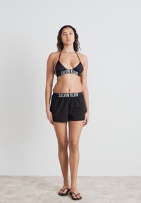 Haut de bikini noir et shorts avec des bandes blanches au logo "Calvin Klein". Le matériau est lisse et ajusté, avec des bretelles réglables sur le haut.