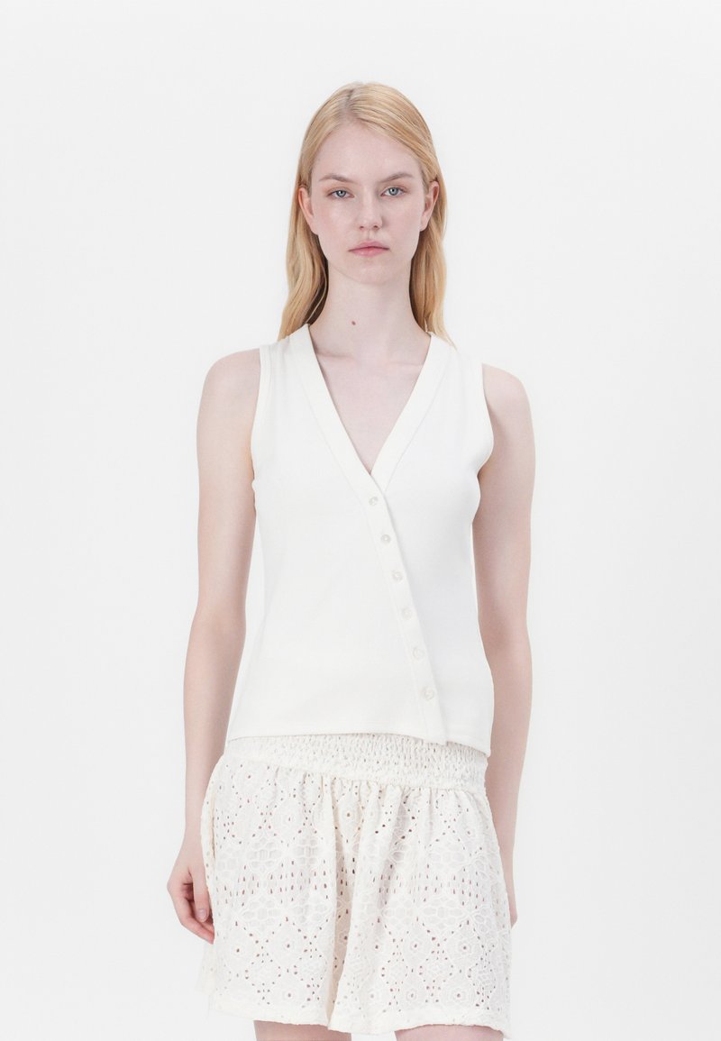 Haut en tricot sans manches blanc avec col en V et patte de boutonnage à six boutons, associé à une jupe blanche à taille élastique brodée. Texture lisse.