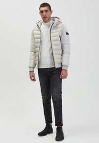 Giacca puffer grigia chiara con cappuccio, chiusura con zip e tasche laterali, indossata sopra un maglione chiaro e jeans scuri, abbinata a stivali neri.
