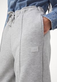 Person trägt hellgraue Jogginghose mit Kordelzugbund und Alpha Industries Logo-Patch, die rechte Hand in der Tasche, sichtbares blaues Jeanshemd.