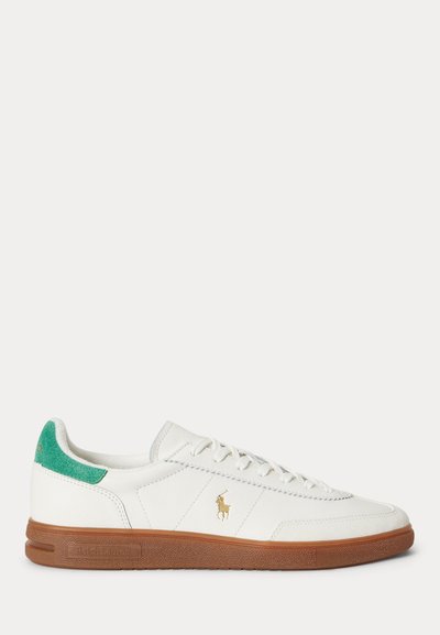 Λευκό sneaker χαμηλής κοπής με πράσινη ταμπέλα στη φτέρνα, καφέ καουτσούκ σόλα, χρυσό λογότυπο Polo στο πλάι και λευκά κορδόνια σε απλό φόντο.