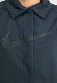 Námornícky modrá bunda Nike s veľkým tmavozeleným vyšívaným logom swoosh a štruktúrovaným golierom, vyrobená z hladkej látky s jemným leskom.