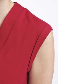 Červený bezrukávový top s výrazným švem na ramene, vyrobený z hladké látky. Má hluboký výstřih do V a čisté okraje.