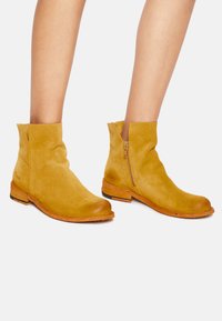 Botas de tobillo de ante en amarillo mostaza con un diseño holgado y cremallera lateral. Suela de madera con ligera textura y puntera redondeada.