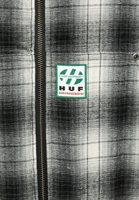 Tissu à carreaux noir et blanc avec une fermeture éclair verticale et un patch bordé de vert portant l'inscription "HUF SAN FRANCISCO 02" en texte vert et rouge.