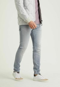 Person som bär en ljusgrå quiltad jacka, lavendelfärgad skjorta, ljusblå slim-fit jeans och vita sneakers med beige och svarta detaljer, stående.