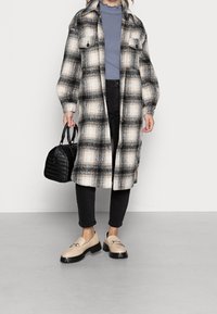 Sort og hvid ternet oversized frakke med to brystlommer, parret med sorte bukser og beige loafers med sorte såler, mens der holdes en lille sort taske.