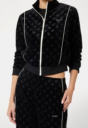 Survêtement en velours noir avec des logos délicats, des détails de passepoil blanc, et la marque "GUESS" sur la poitrine de la veste et la cuisse du pantalon.