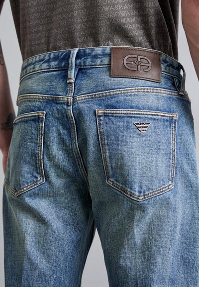 Light blue denim jeans feature a brown leather waistband patch with logo, visible stitching, and two back pockets with logo detail.
