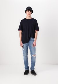 Marineblå kortærmet t-shirt med røde og hvide detaljer, sammen med lyseblå slidte jeans og sorte sneakers, iført en sort bøttehat.