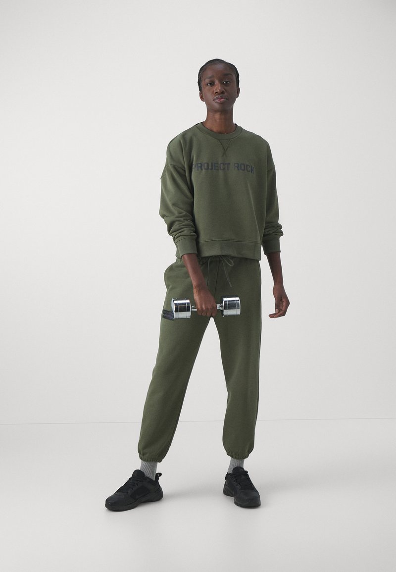 Olivgrüner, verkürzter Sweatshirt mit dem Text "PROJECT ROCK", passenden Jogginghosen und schwarzen Sneakern, hält ein silbernes Hantel.