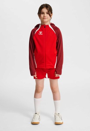 Ung pige stående iført en rød og mørkerød hættetrøje med lynlås, matchende røde shorts, hvide crew-sokker og hvide sneakers på en ensfarvet baggrund.