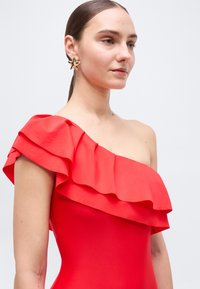 Γυναίκα με κόκκινο φόρεμα ενός ώμου με λεπτομέρεια βολάν, που φοράει χρυσά σκουλαρίκια σε σχήμα αστεριού, σε απλό ανοιχτό φόντο.