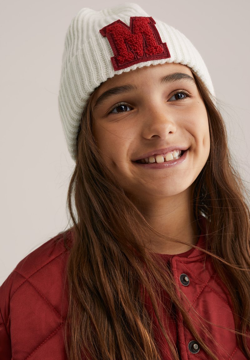 Baltas ribuotas megztas bžūpė su raudonu pūkuotu „M“ lopinėliu priekyje. Fonas neutralus, išryškinantis kepurės dizaino detales.