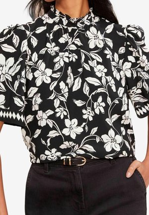 Mujer que lleva una blusa negra con mangas abullonadas y estampado floral blanco combinada con pantalones negros y un cinturón negro con hebilla dorada.