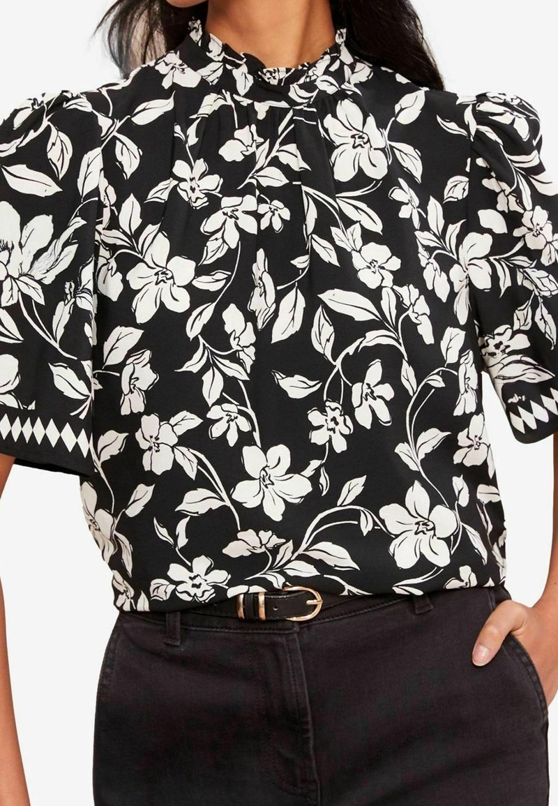 Femme portant un chemisier noir à manches bouffantes avec imprimé floral blanc, associé à un pantalon noir et une ceinture noire avec boucle dorée.