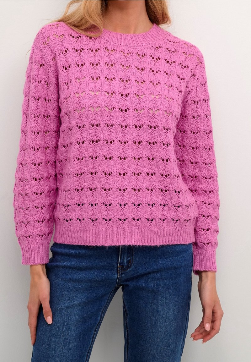 Gestrickter rosa Pullover mit einem strukturierten, offenen Muster, runden Ausschnitt und gerippten Bündchen und Saum. Kombiniert mit blauen Jeans.