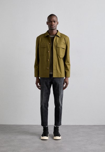 Grøn bomuldsjakke med to brystlommer, sort t-shirt under, grå denimjeans og sorte sneakers med høj skaft og hvide såler.