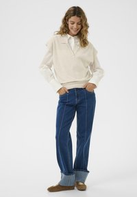 Beige kötött ujjatlan felső egy fehér ing fölött, párosítva kék farmernadrággal, oldalsó varrással és bővülő szárakkal. Barna cipők egészítik ki a szettet.