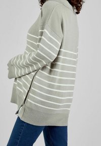 Femme portant un pull ample gris clair avec des rayures blanches horizontales et un jean bleu, vue de profil sur un fond uni.