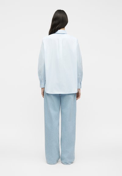 Γυναίκα με μακριά σκούρα μαλλιά, ντυμένη με ανοιχτό μπλε πουκάμισο με κουμπιά και φαρδιά ανοιχτά τζιν παντελόνια, στέκεται με την πλάτη προς ένα απλό φόντο.