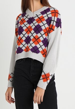 Kobieta ubrana w krótki sweter z wzorem w romby w kolorach fioletowym, pomarańczowym i białym na klatce piersiowej i rękawach, w połączeniu z czarnymi spodniami z wysokim stanem.